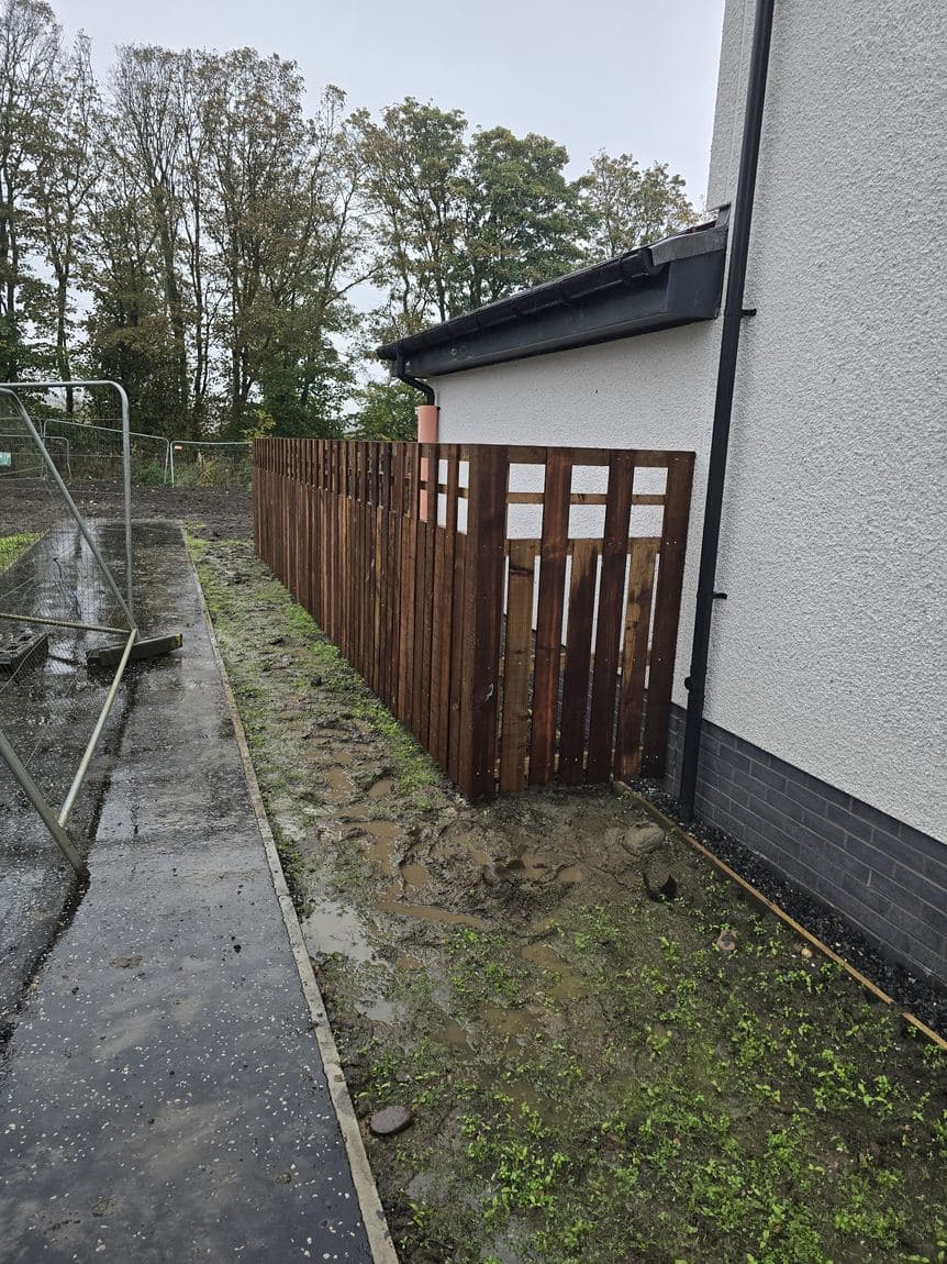 Short vertical-board timber fence against a house wall in East Calder